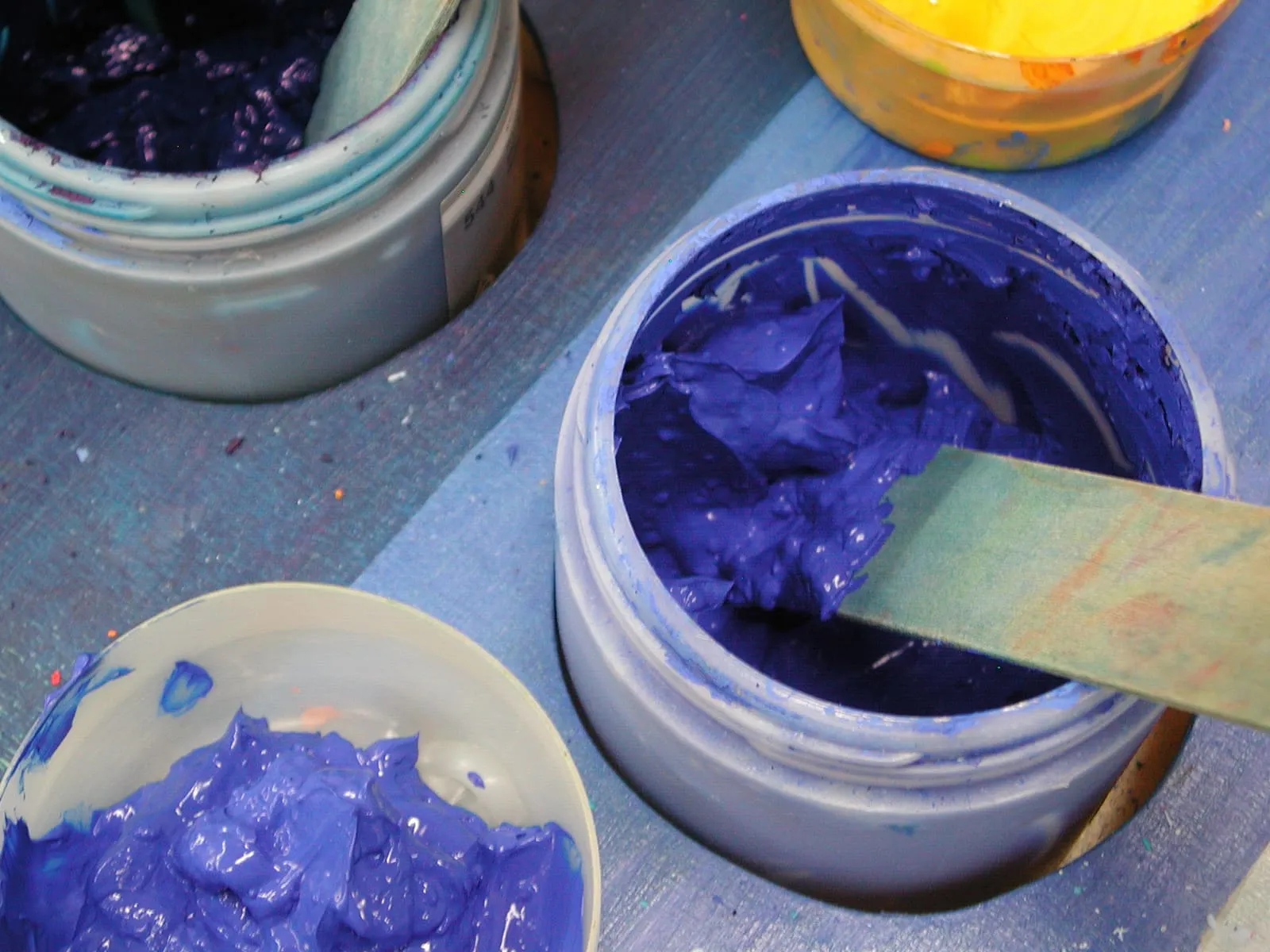 Drei offene Farbtöpfe mit dicker blauer, violetter und gelber Farbe auf einem bemalten Brett, darin ein Spachtel.